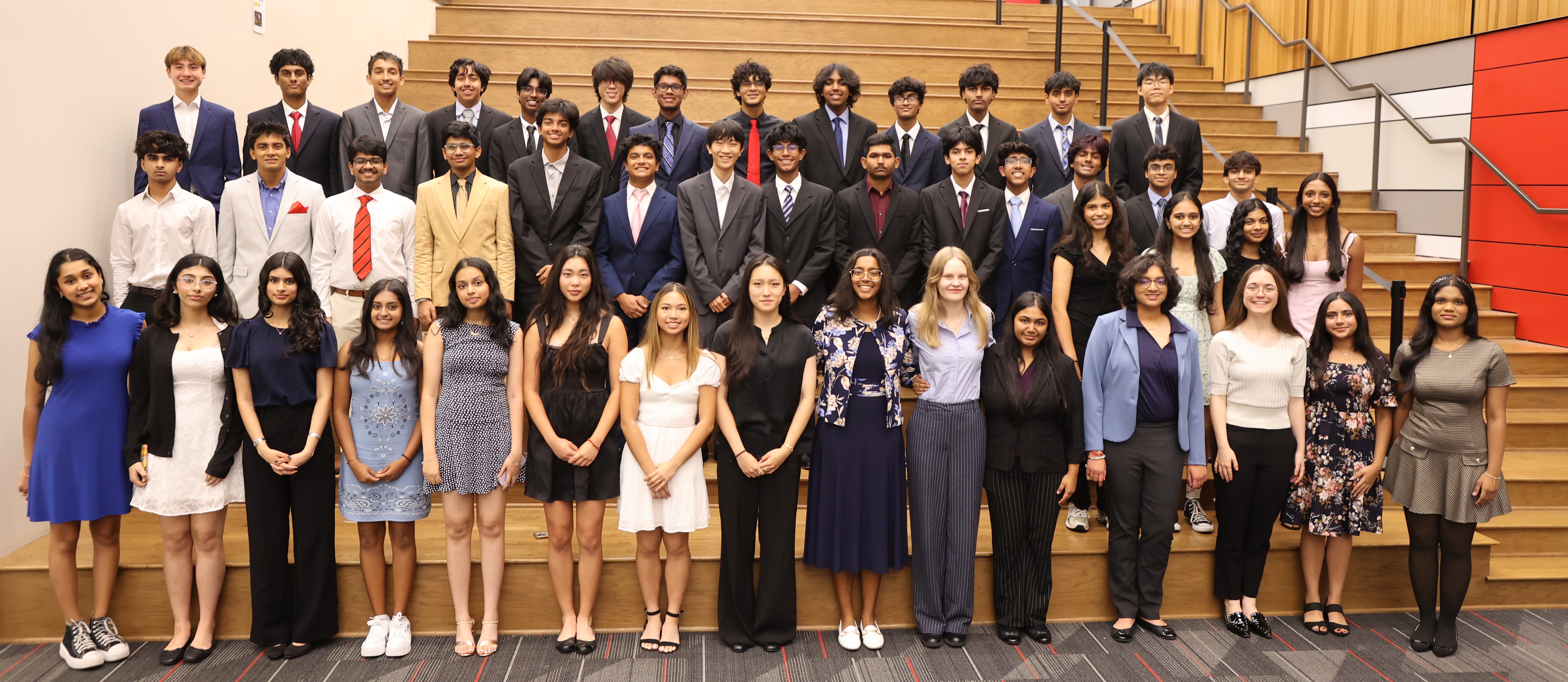 Group photo of National Merit Semifinalists from Coppell High School, posed in three rows on a wooden amphitheater-style staircase beneath the red and white Coppell sign. About 45 students stand together in formal attire, with the boys in suits and ties in the back and middle rows, and the girls in dresses and blouses in the front row.