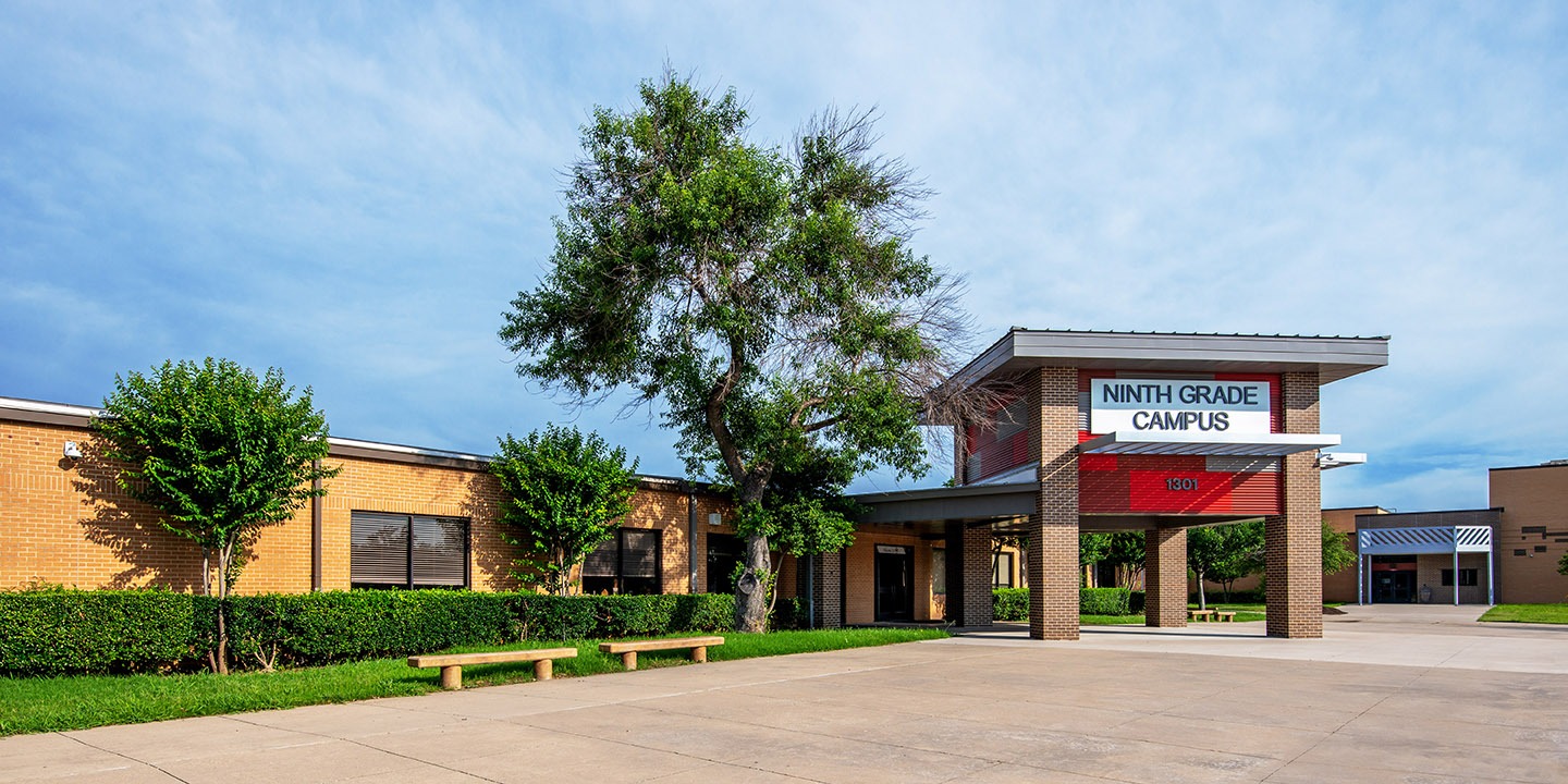 A single-story brick school building with a covered entrance labeled "Ninth Grade Campus," trees, hedges, and benches in the foreground.