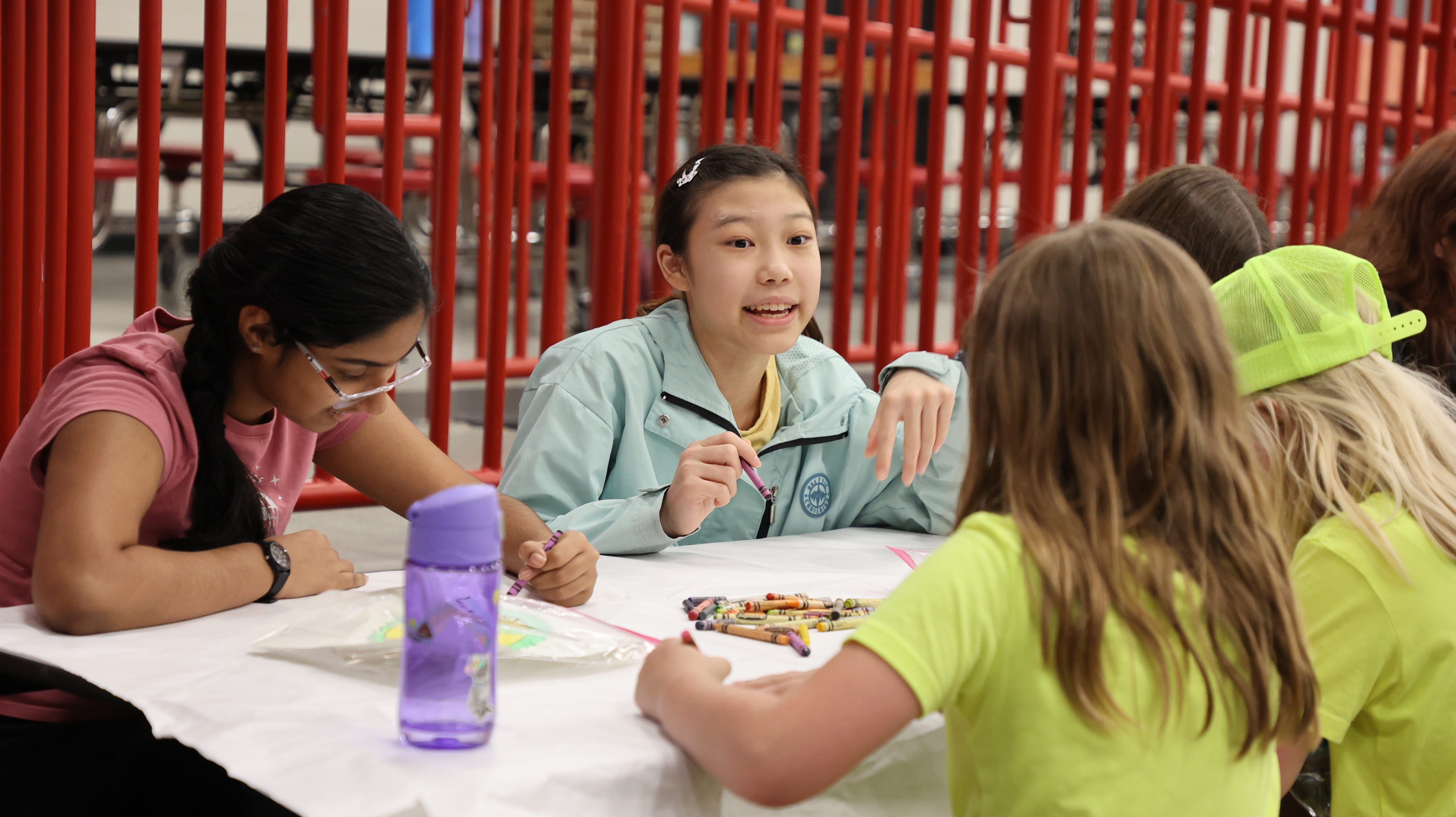 Students sit together at a table coloring with crayons on a paper-covered surface in a school cafeteria.