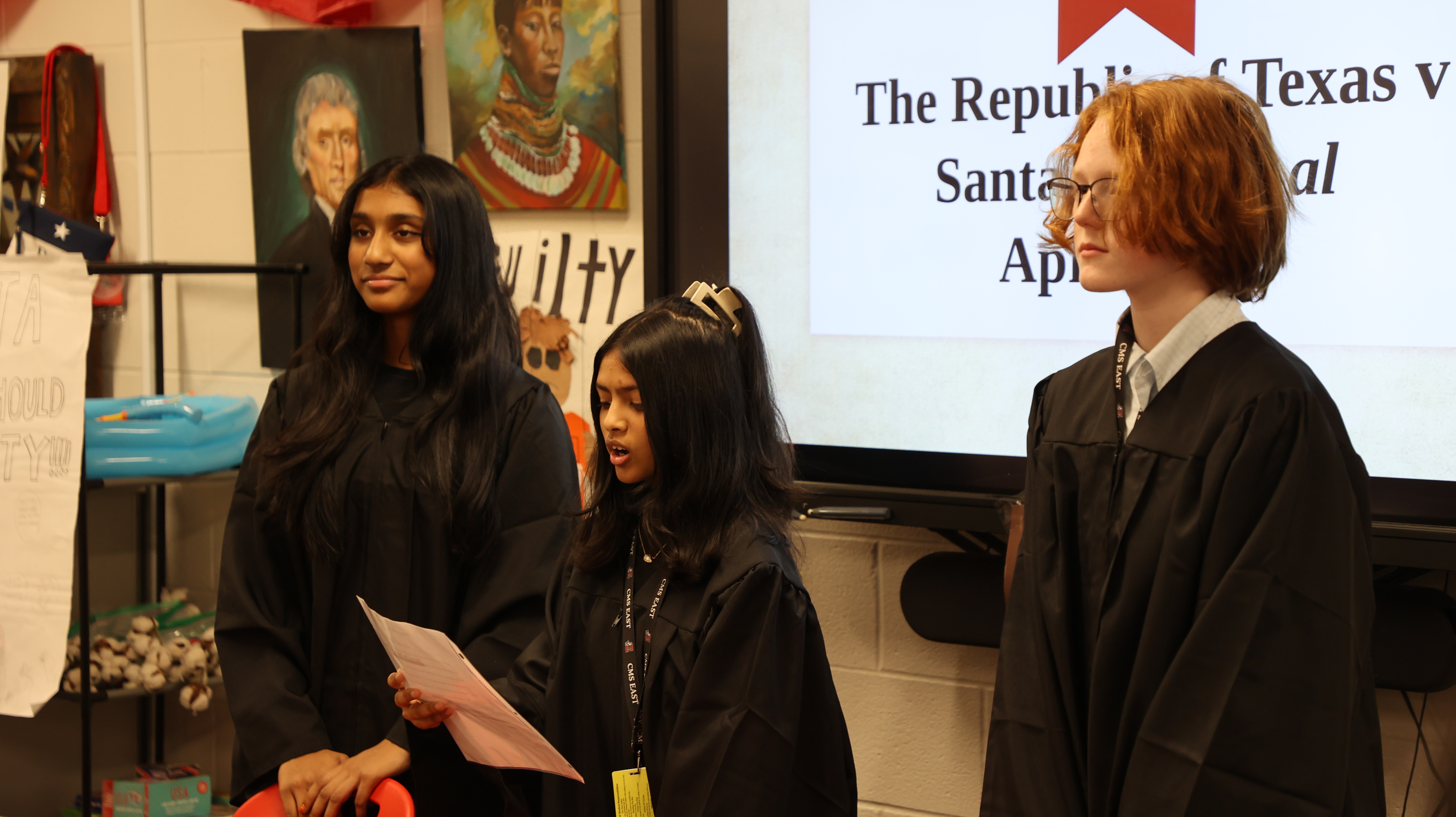 Three middle school students in black graduation-style robes stand in a classroom, presenting a mock trial titled "The Republic of Texas v. Santa Anna" displayed on a screen behind them, with classroom artwork and historical portraits on the wall.
