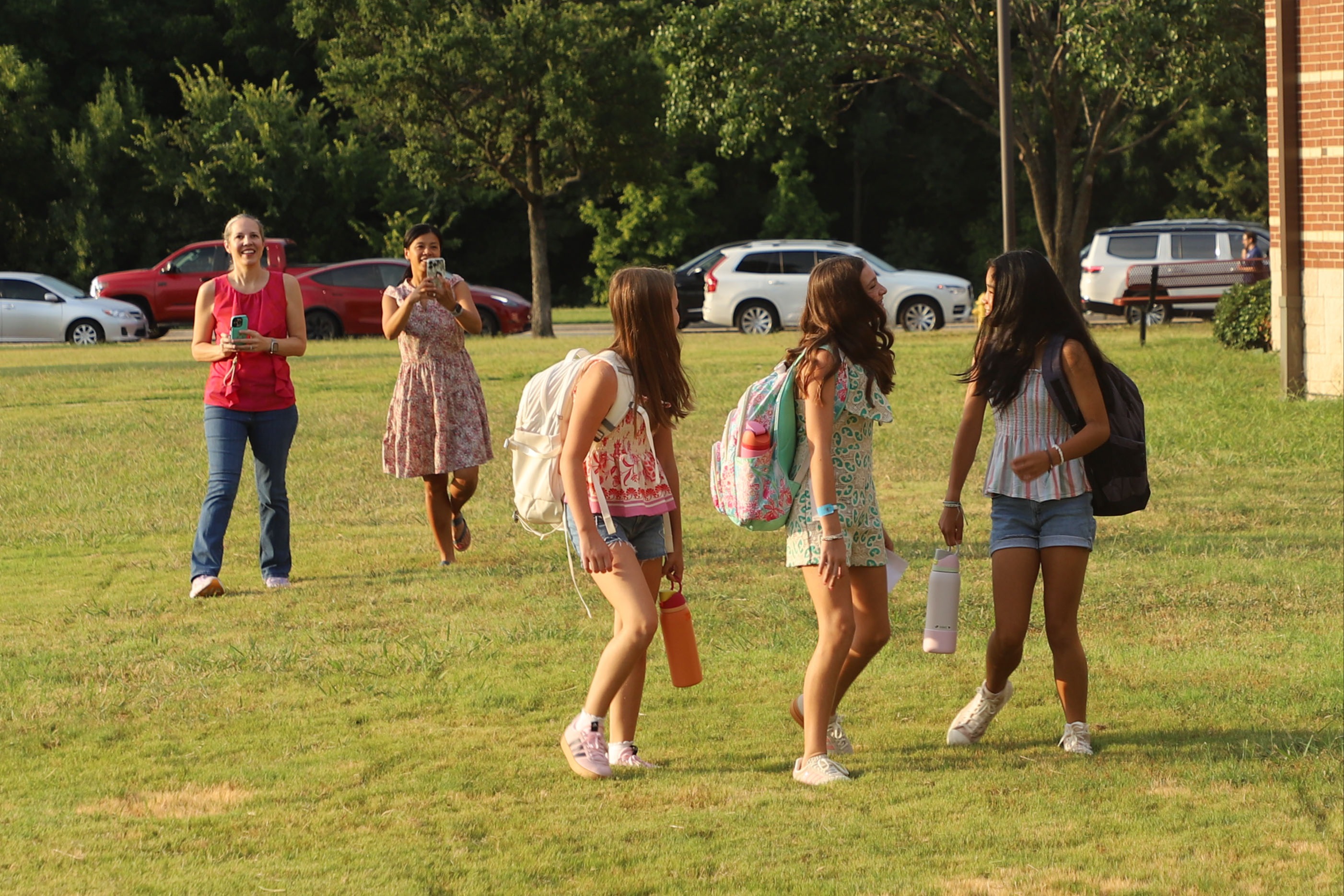 Three girls with backpacks walk across a grassy schoolyard while two parents take photos of them on the first day of school.