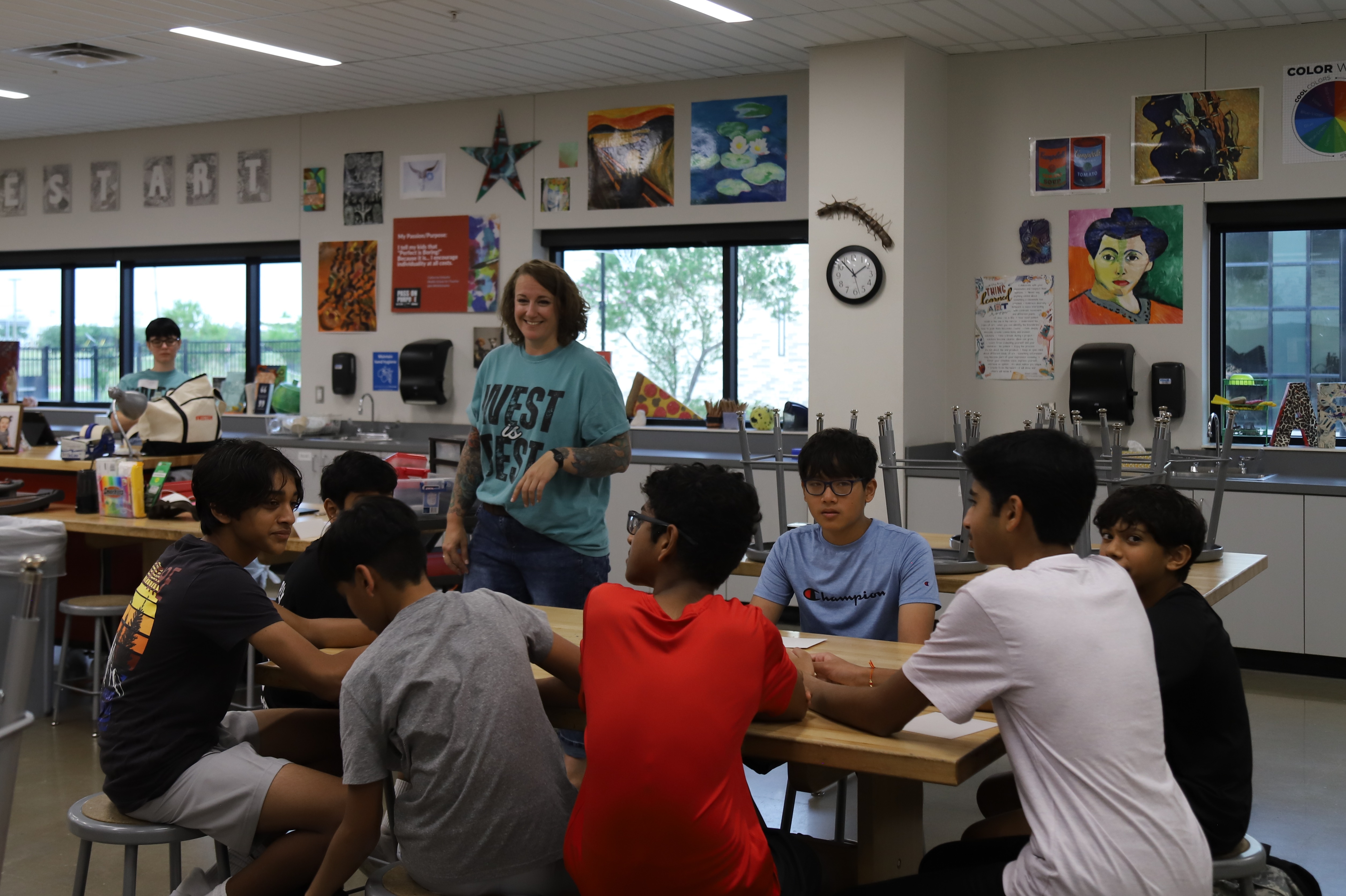 A teacher smiles while standing near a group of students seated around a table in an art classroom decorated with student artwork.