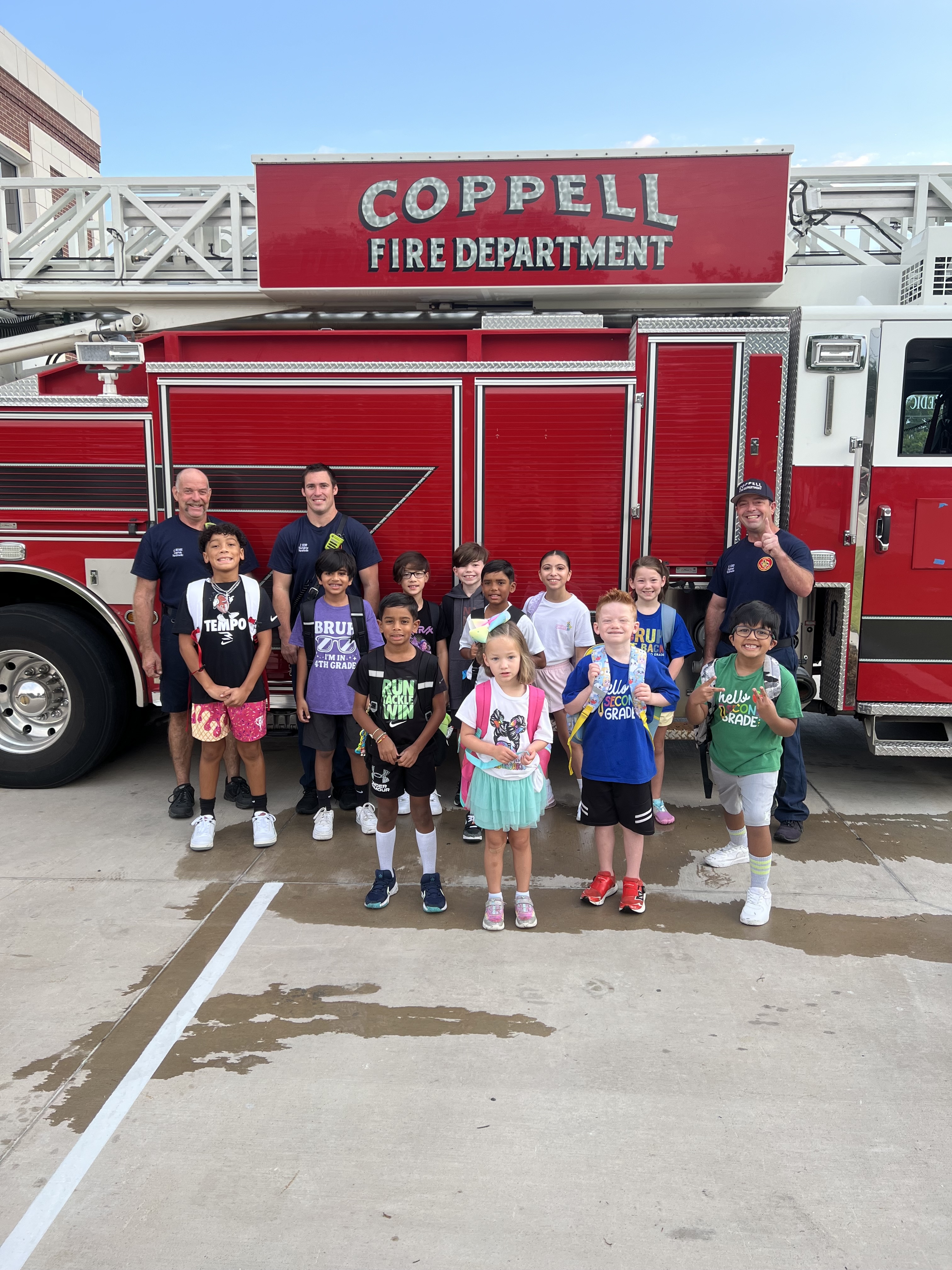 A group of elementary school children and firefighters pose smiling in front of a red Coppell Fire Department ladder truck outside the fire station.