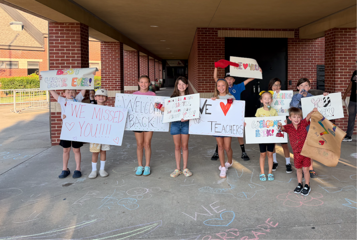 A group of smiling children hold handmade "Welcome Back" signs for teachers outside a school entrance decorated with sidewalk chalk art.