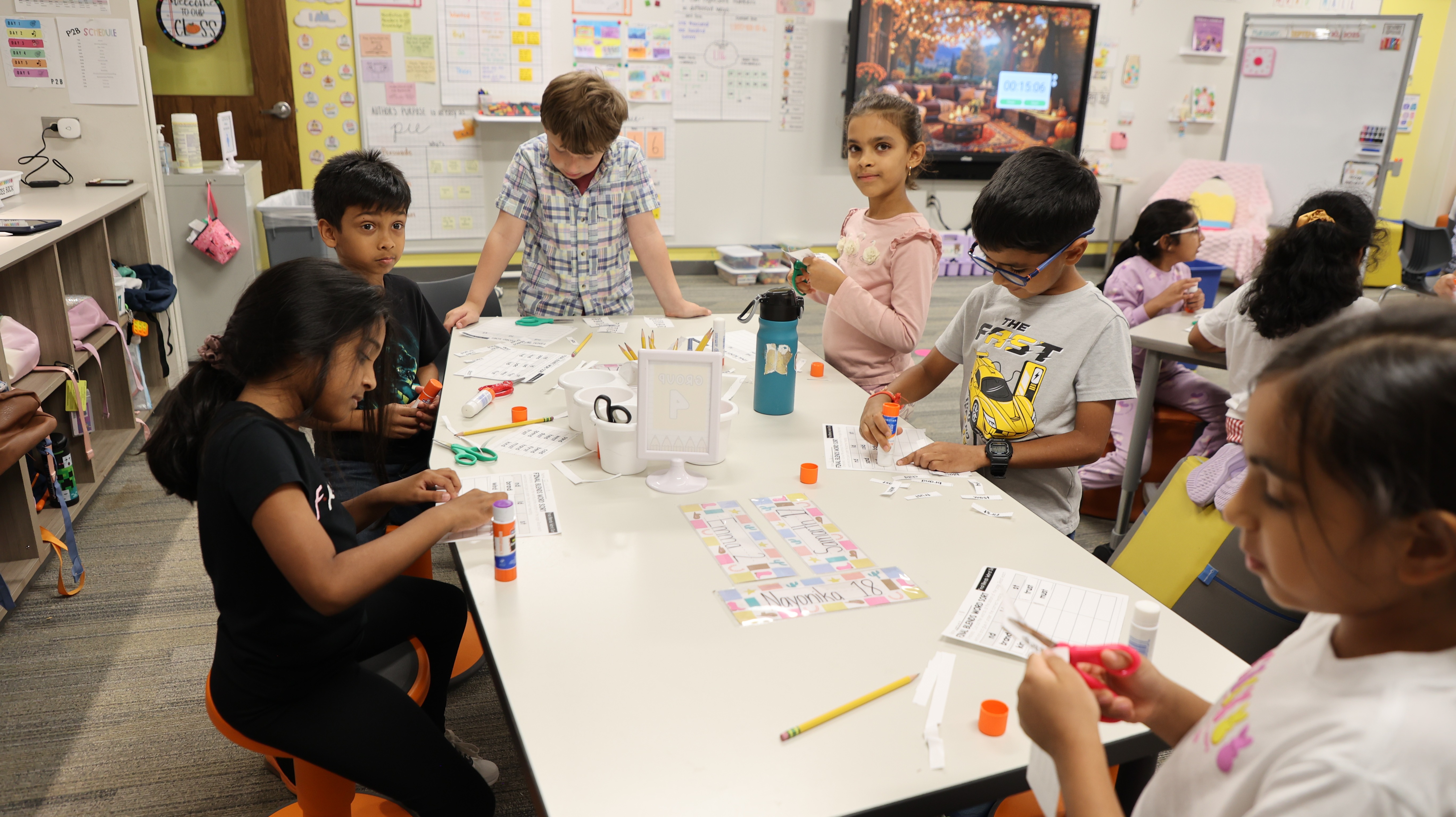 A group of elementary students works together at a table using scissors, glue sticks, and word cards for a classroom activity.