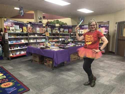blond woman dressed in a tutu stands in front of tables and shelves of books