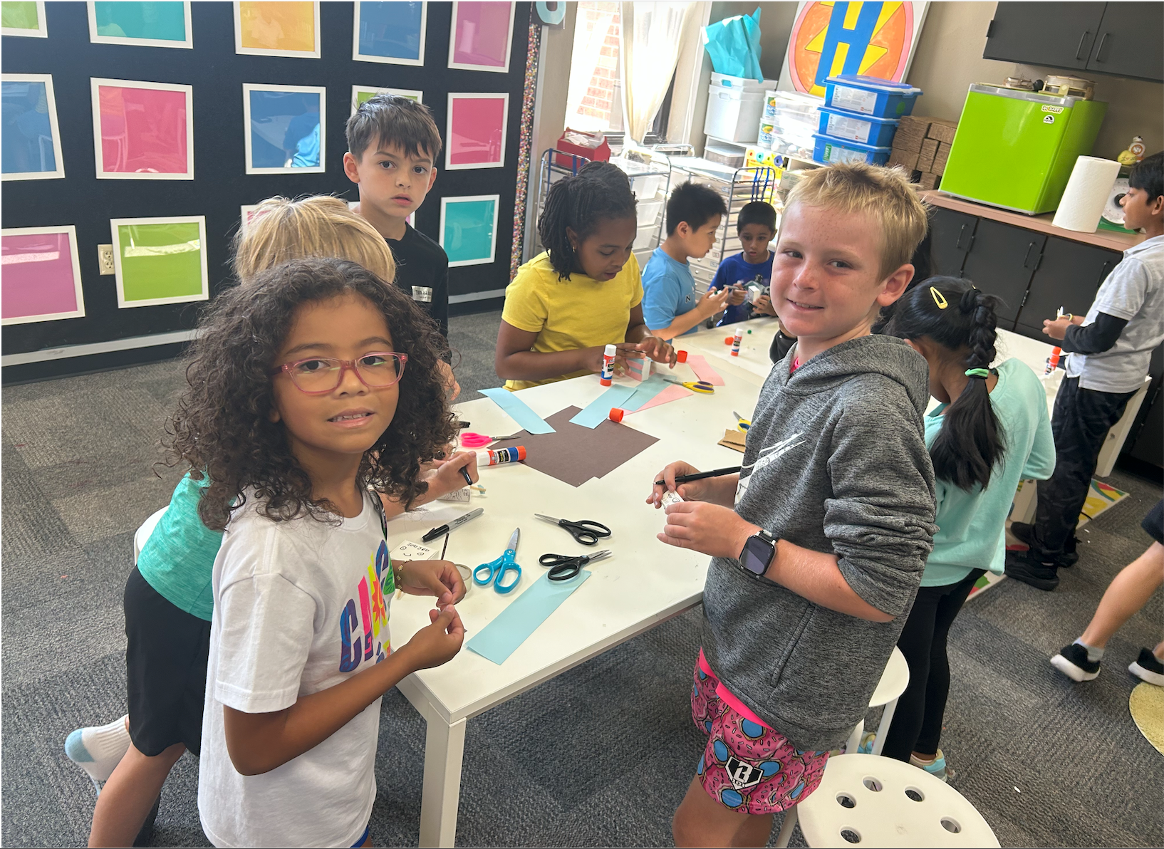 Children work on a craft project with construction paper, scissors, and glue sticks at a table in a colorful classroom.