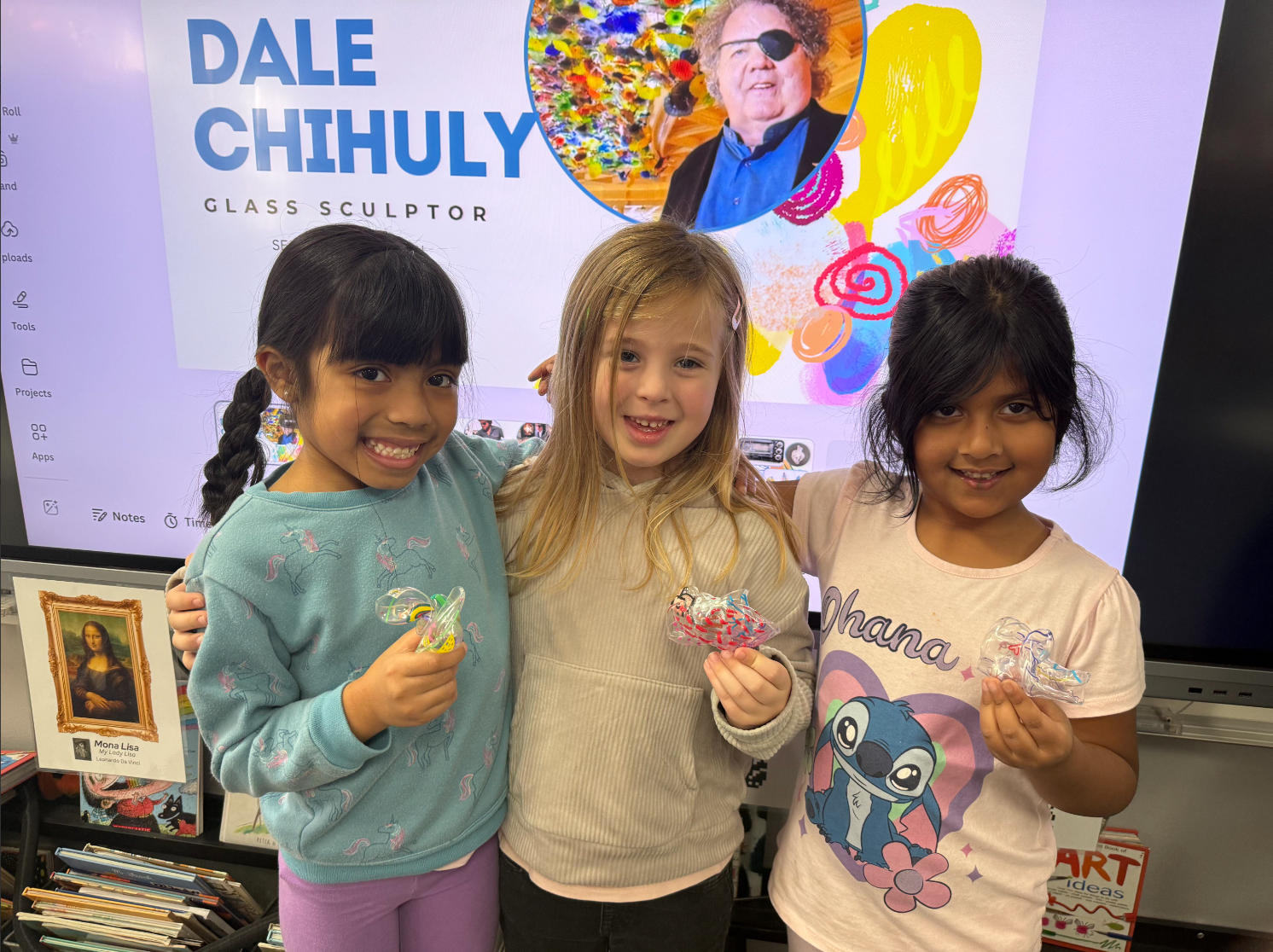 Three young girls smile with their arms around each other, each holding a small handmade glass or plastic art piece. Behind them, a classroom screen displays a lesson on glass sculptor Dale Chihuly, and a Mona Lisa print sits on a shelf to the left.