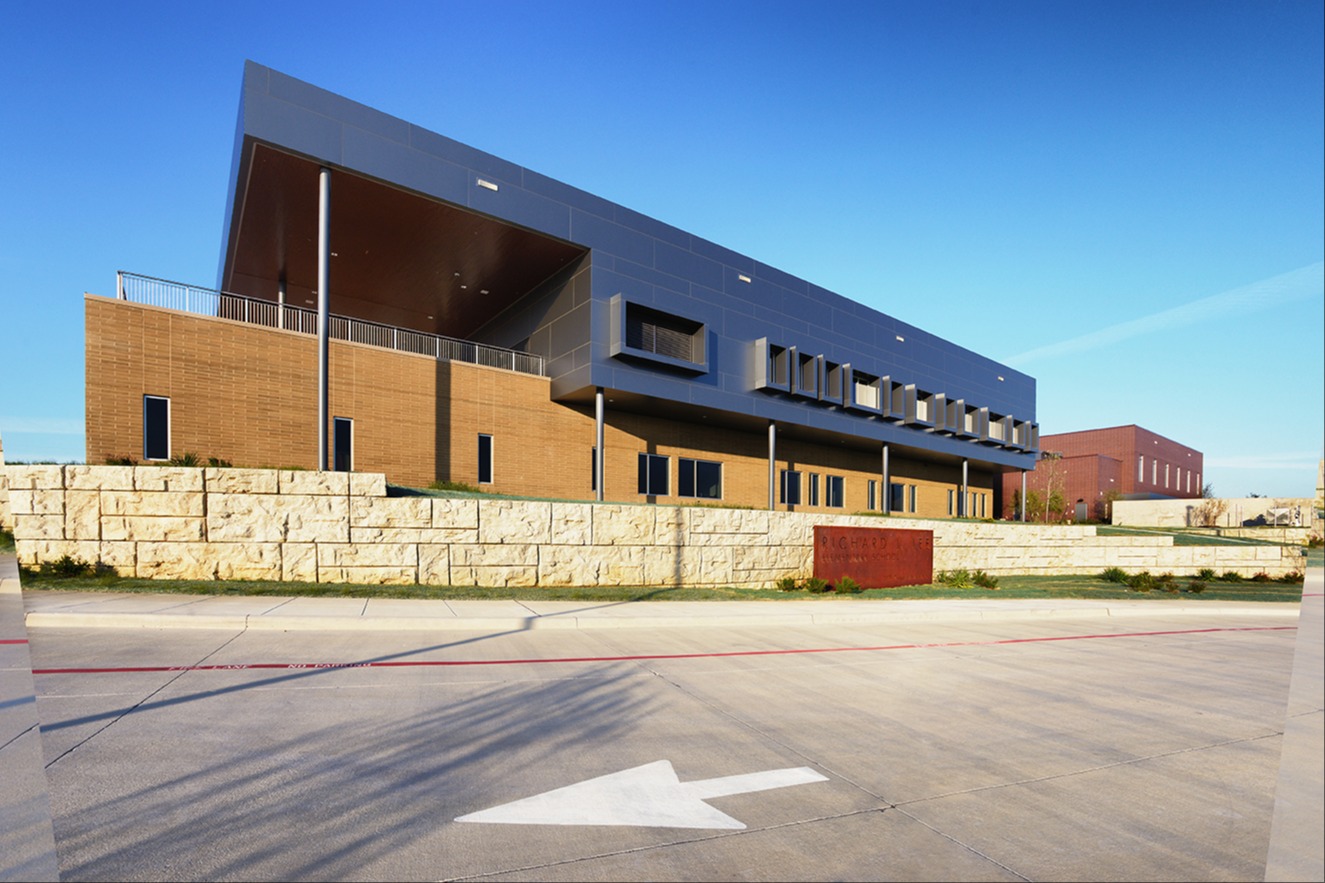 Modern school building with an angled metal roof, brick lower level, and a stone retaining wall bearing a "Richard Lee Elementary School" sign in front.
