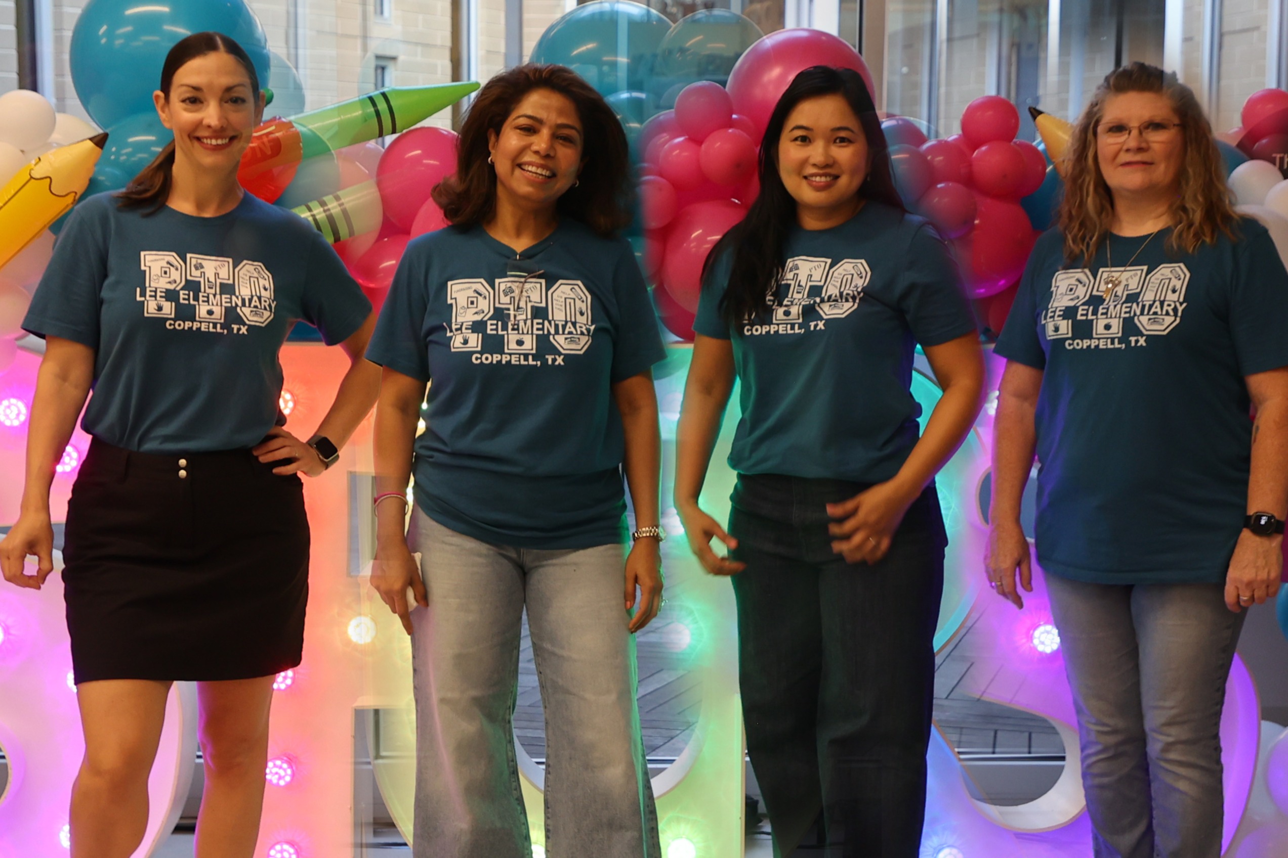 Four women stand together wearing matching teal "PTO Lee Elementary, Coppell, TX" t-shirts, smiling in front of a colorful balloon backdrop with giant pencil and crayon decorations and illuminated letters.