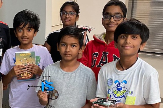 A group of students pose indoors, several holding LEGO robotics creations and one holding a book, smiling together in a classroom setting.