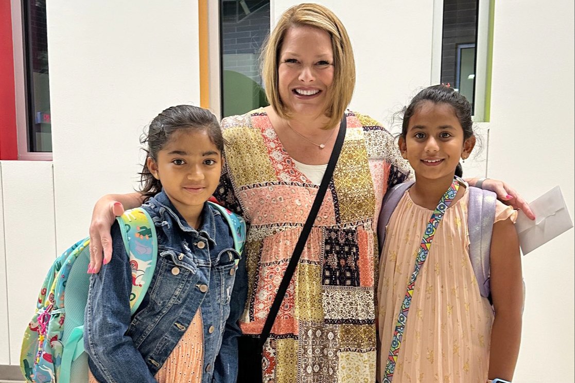 A smiling woman in a colorful patterned dress poses with her arms around two young girls wearing backpacks, standing indoors near large windows.