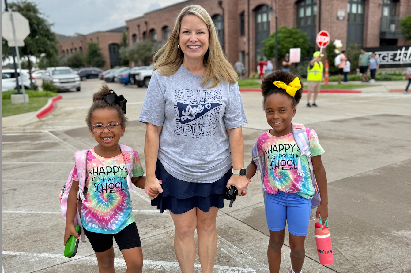 A smiling adult holds hands with two young students outside a brick school building. The students wear colorful “Happy Last Day of School” shirts and backpacks, while the adult wears a gray Lee Spurs T-shirt.