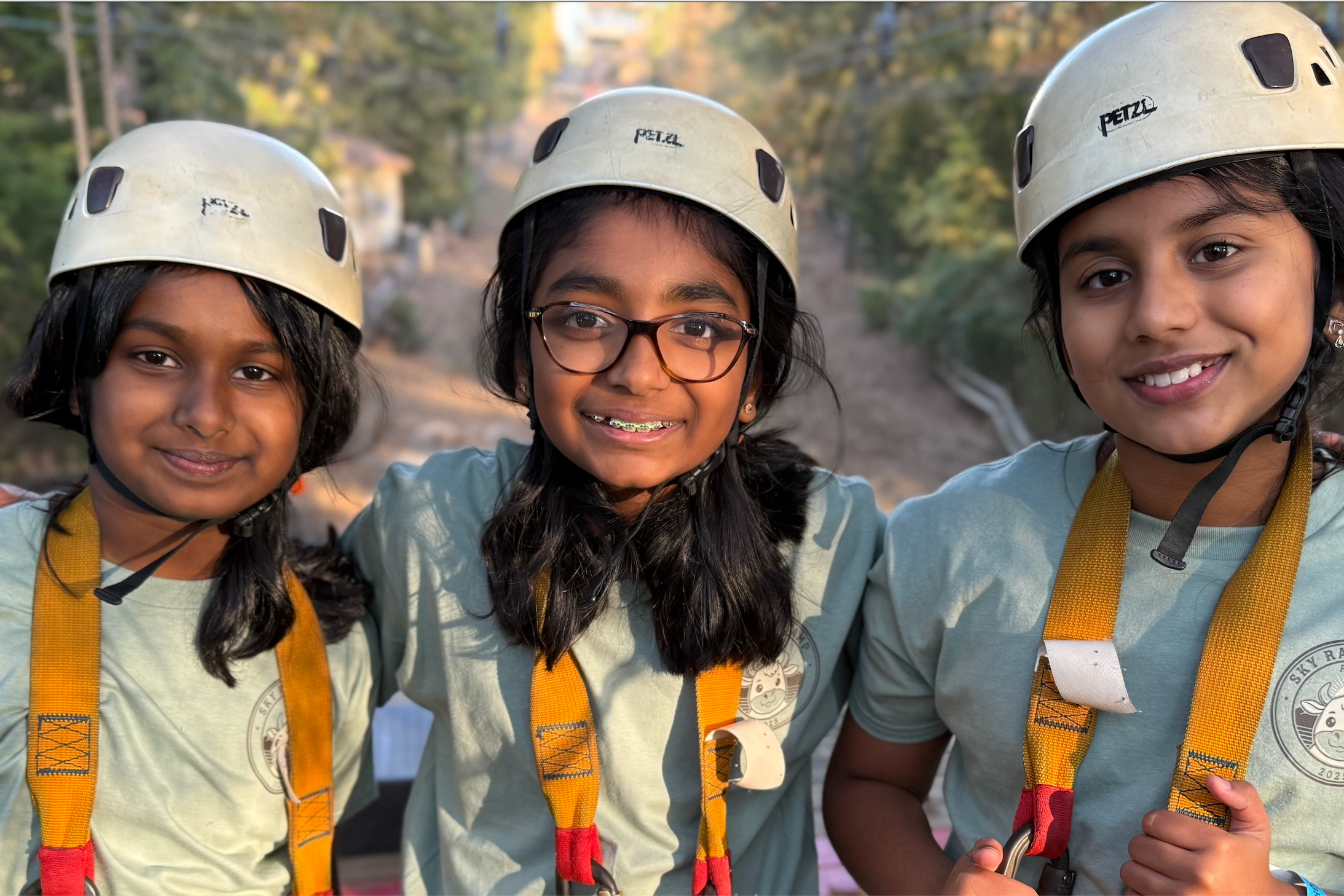 Three smiling students wearing helmets, safety harnesses, and matching camp T-shirts stand with their arms around one another at an outdoor ropes course surrounded by trees.