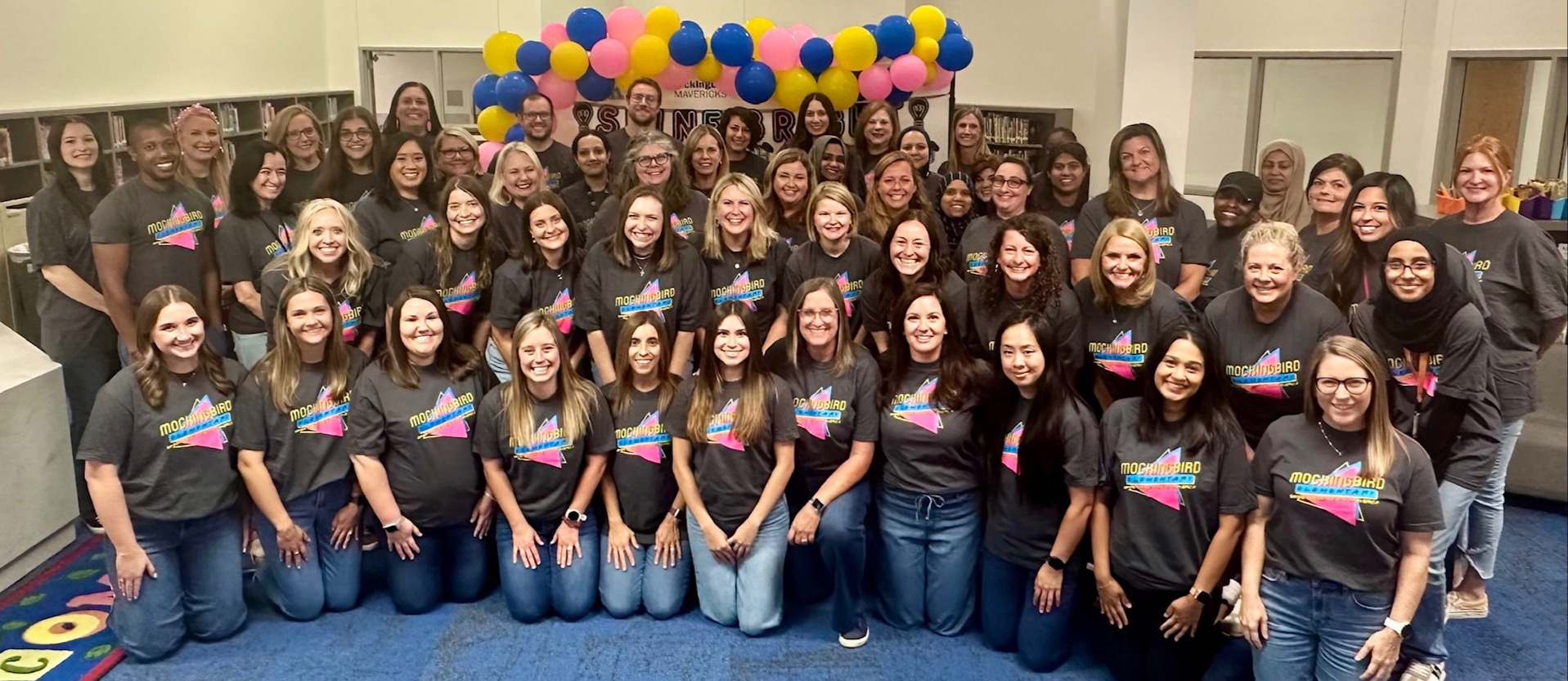 This is a picture of the staff of Mockingbird Elementary for the 2025-2026 school year. The kneeling and standing in rows in the school library and all wearing matching t-shirts.