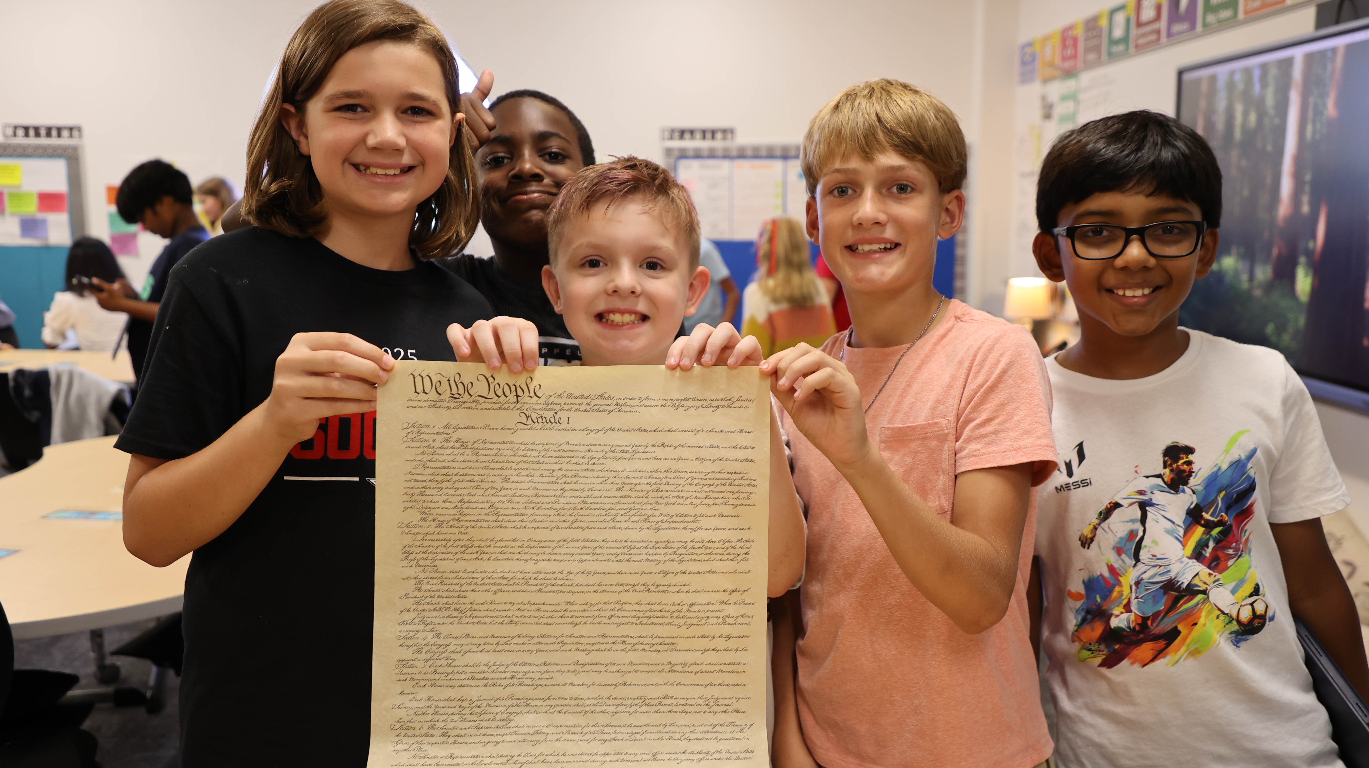 Five students smile in a classroom while holding up a reproduction of the "We the People" Constitution document.