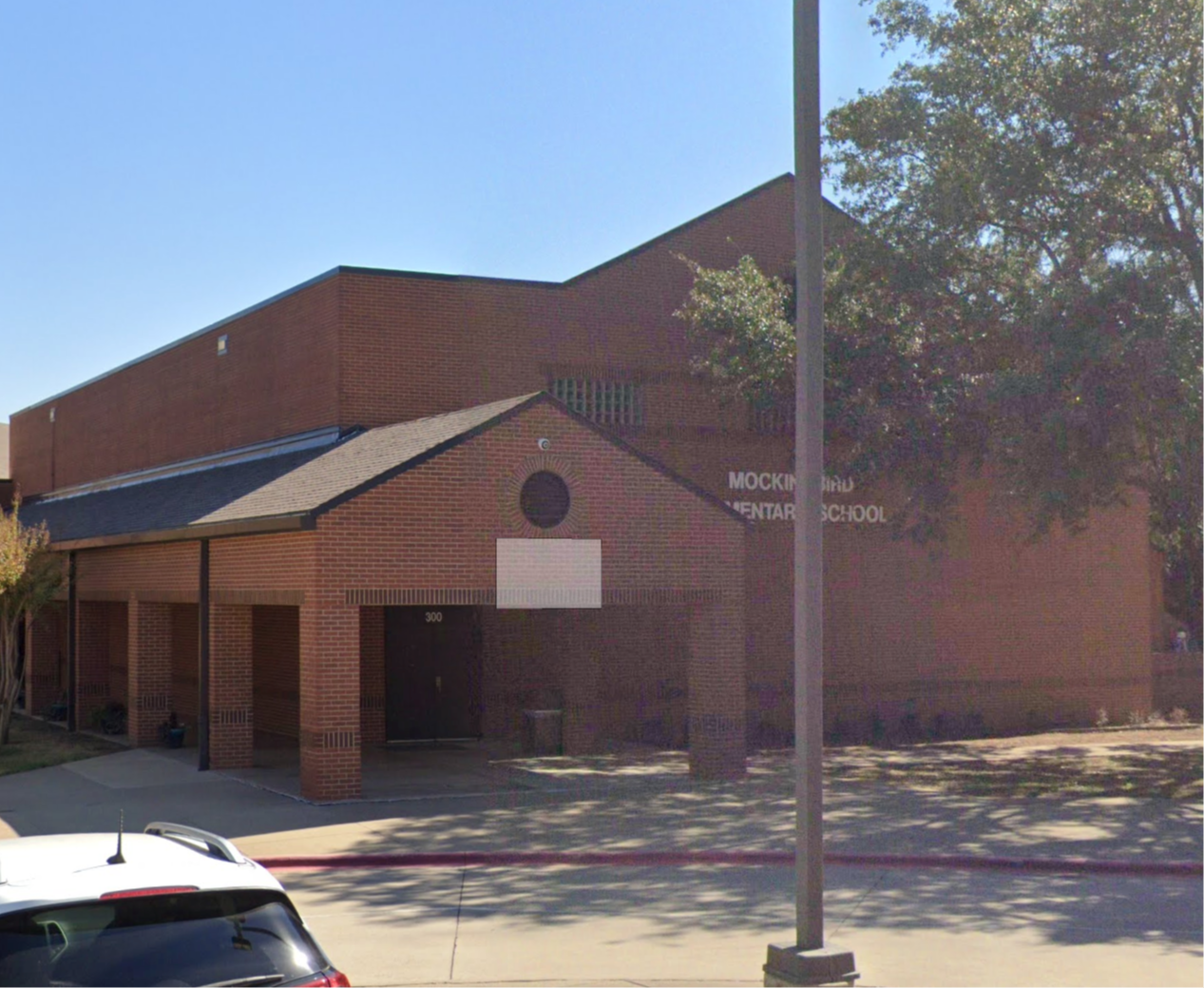 Brick building labeled "Mockingbird Elementary School" with a covered entrance walkway, trees, and a parked car in front.