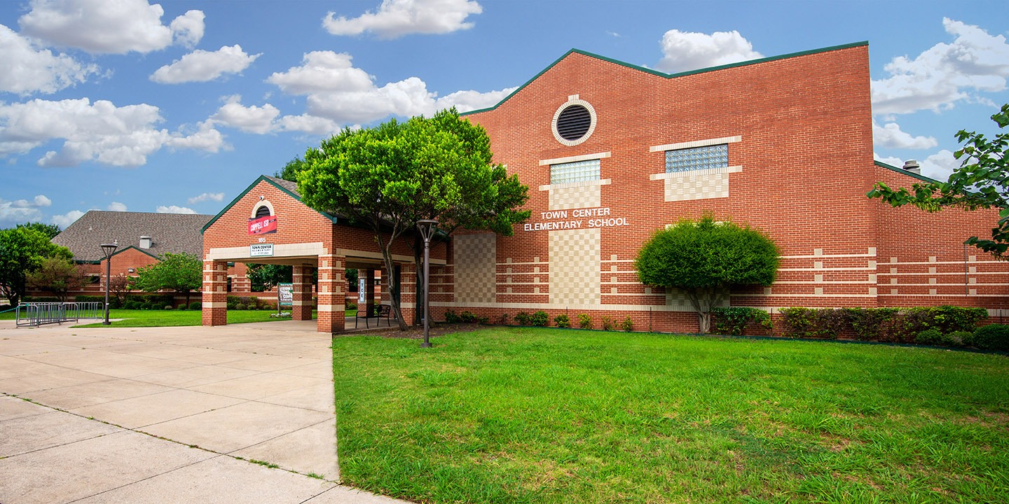 Brick building labeled "Town Center Elementary School" with a covered entrance, tree-lined lawn, and a partly cloudy sky.