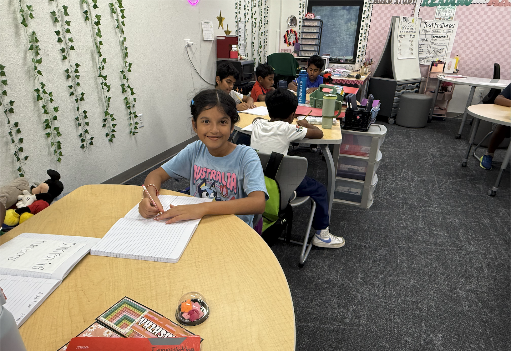 A young girl in an "Australia" T-shirt smiles at the camera while writing in a notebook at a classroom table, with other students working at desks in the background.