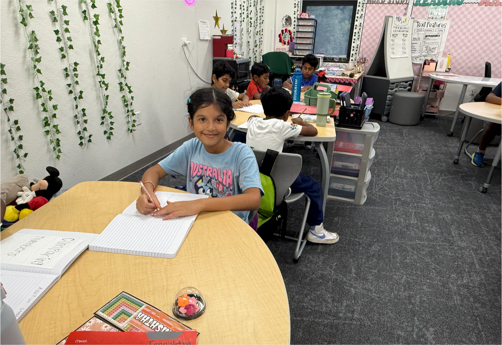 A girl in an "Australia" t-shirt smiles at the camera while writing in a notebook at a round table, with other students working at desks in the background of a classroom decorated with hanging vine plants and reading posters.