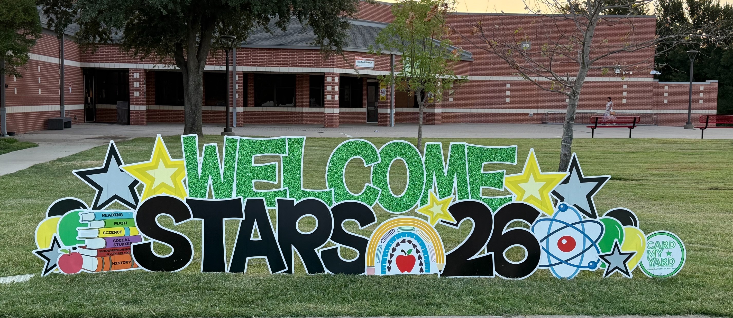 Sign staked in front of school with text: Welcome Stars 26 and star graphics, and other back to school graphics