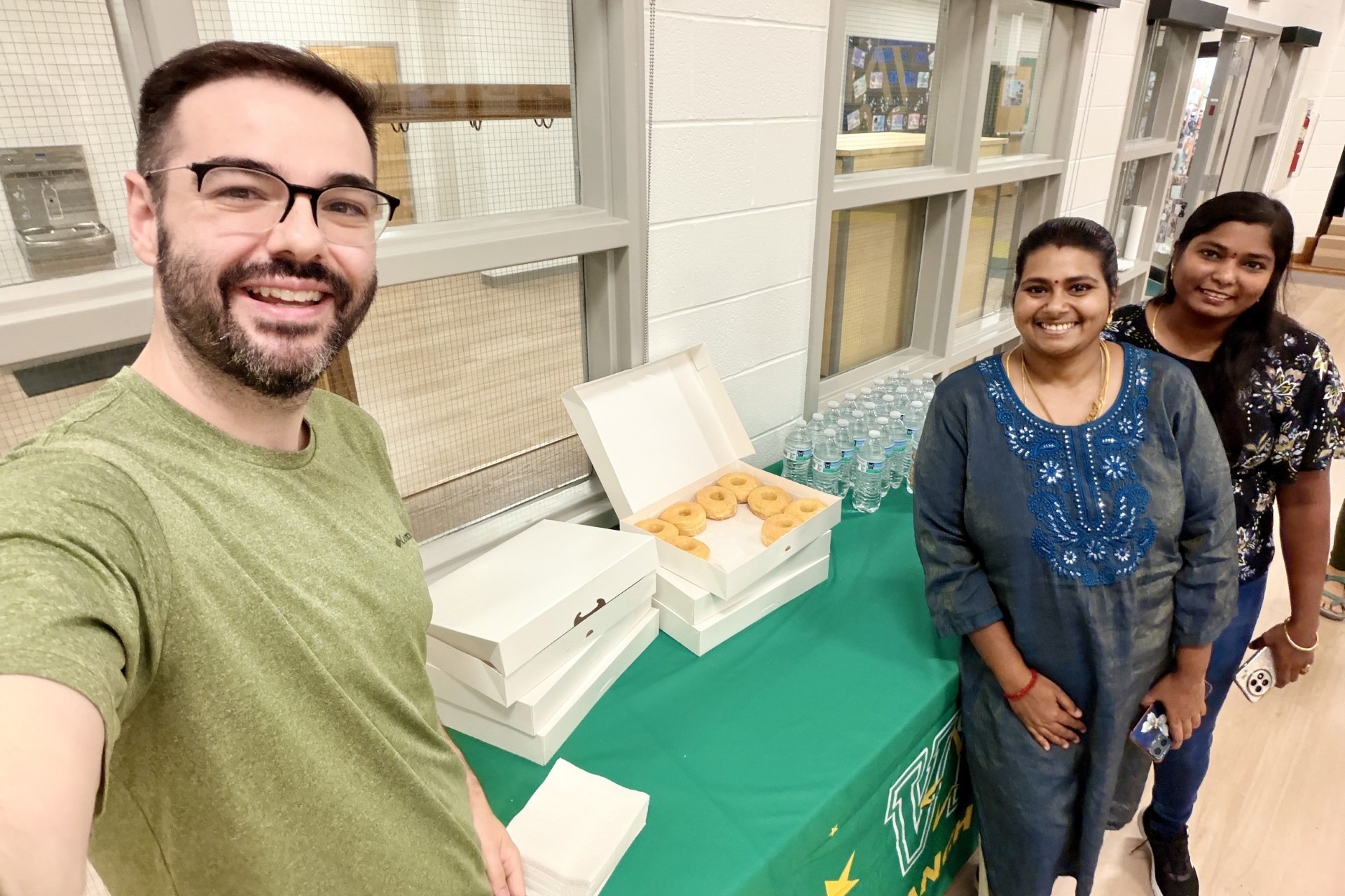 Group of three smiling adults standing around a table covered with a green tablecloth. On the table are bottles of water and boxes of donuts.