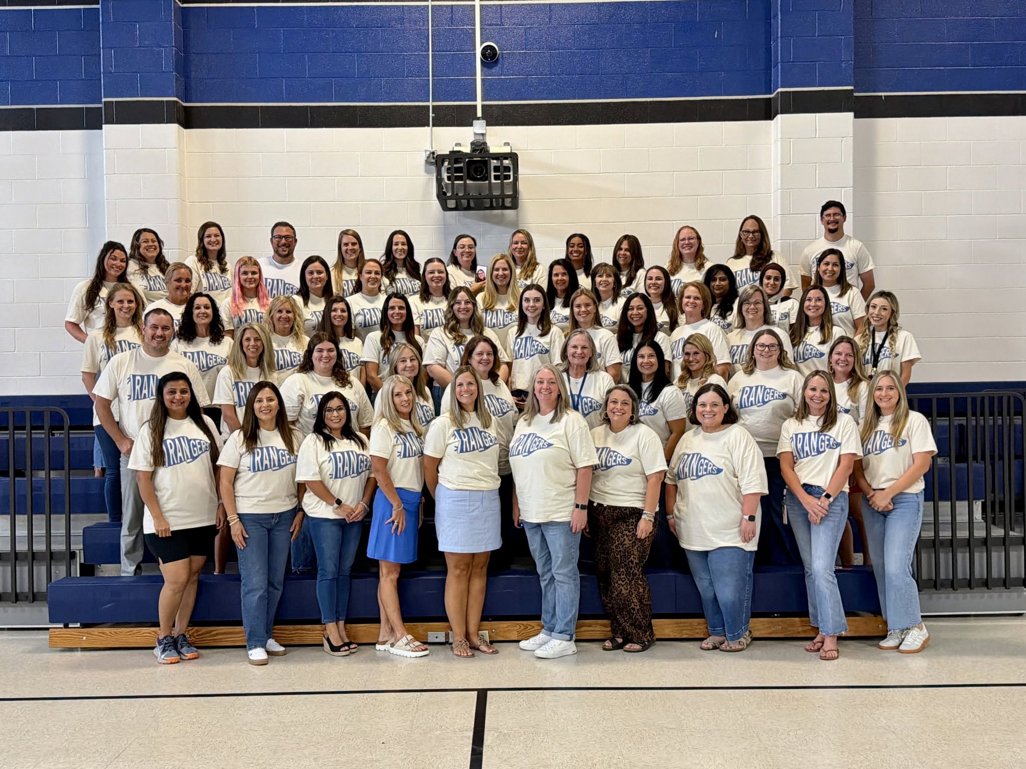 A large group photo of school staff wearing matching cream "Rangers" T-shirts, standing in rows on bleachers in a gymnasium.