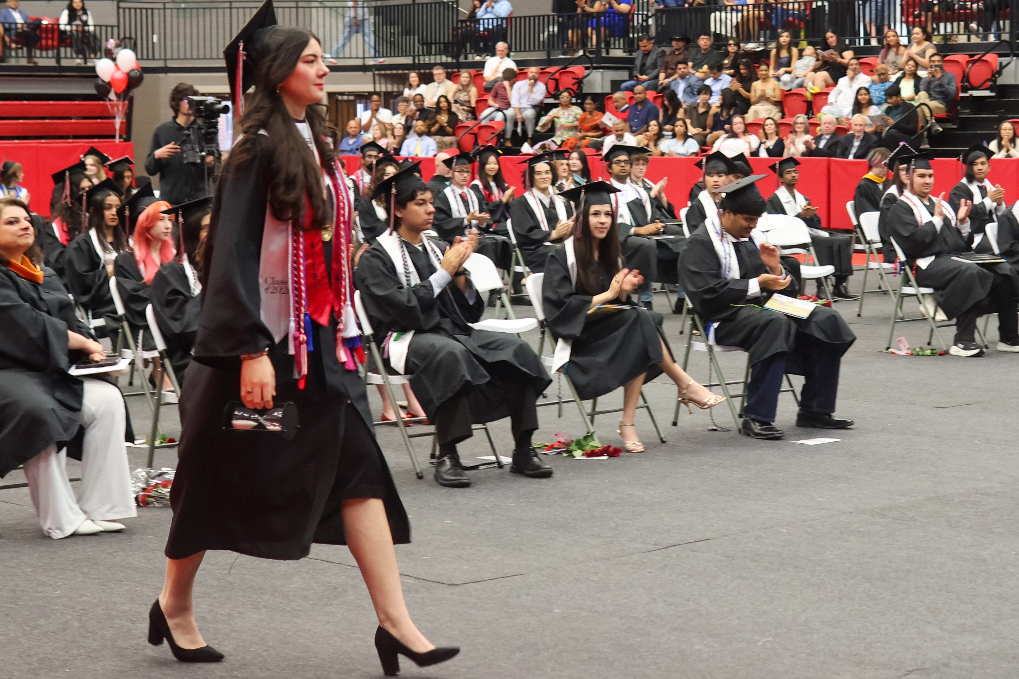 A graduate in a cap and gown with honor cords walks across the floor while seated classmates applaud during an indoor graduation ceremony.