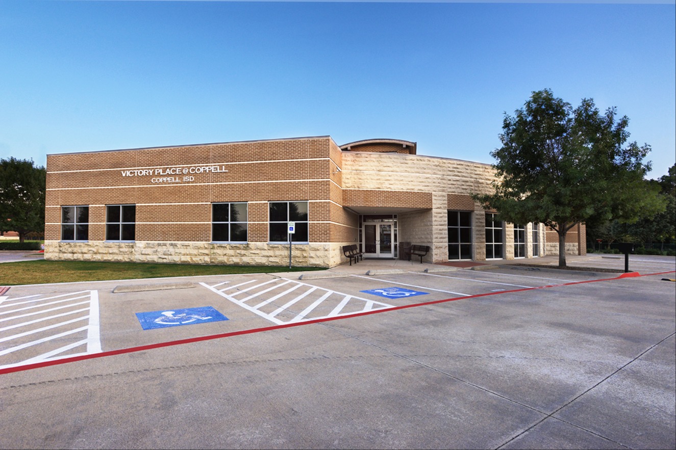 Brick building labeled "Victory Place @ Coppell, Coppell ISD" with a front entrance and parking lot in the foreground.