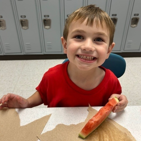 A young child with short brown hair, wearing a red shirt, smiles brightly while holding and eating a slice of watermelon in a school cafeteria with lockers in the background.