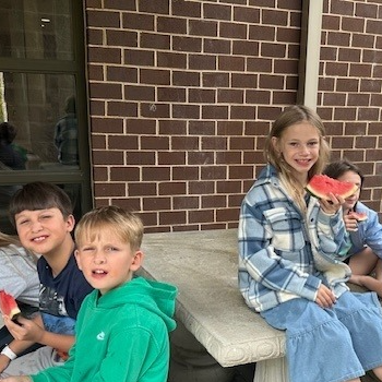 Several young children eating watermelon sitting on a table with a brick wall in the background. 