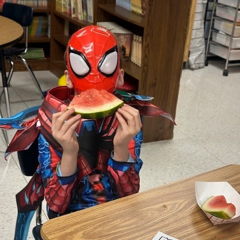 A child dressed in a Spider-Man costume and mask is sitting in a classroom or library setting, holding and eating a slice of watermelon. Bookshelves and a table are visible in the background.