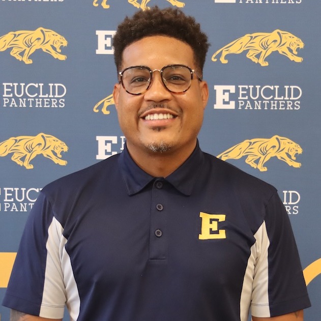 A man with glasses and a tattoo stands in front of a wall with blue and yellow graphics.