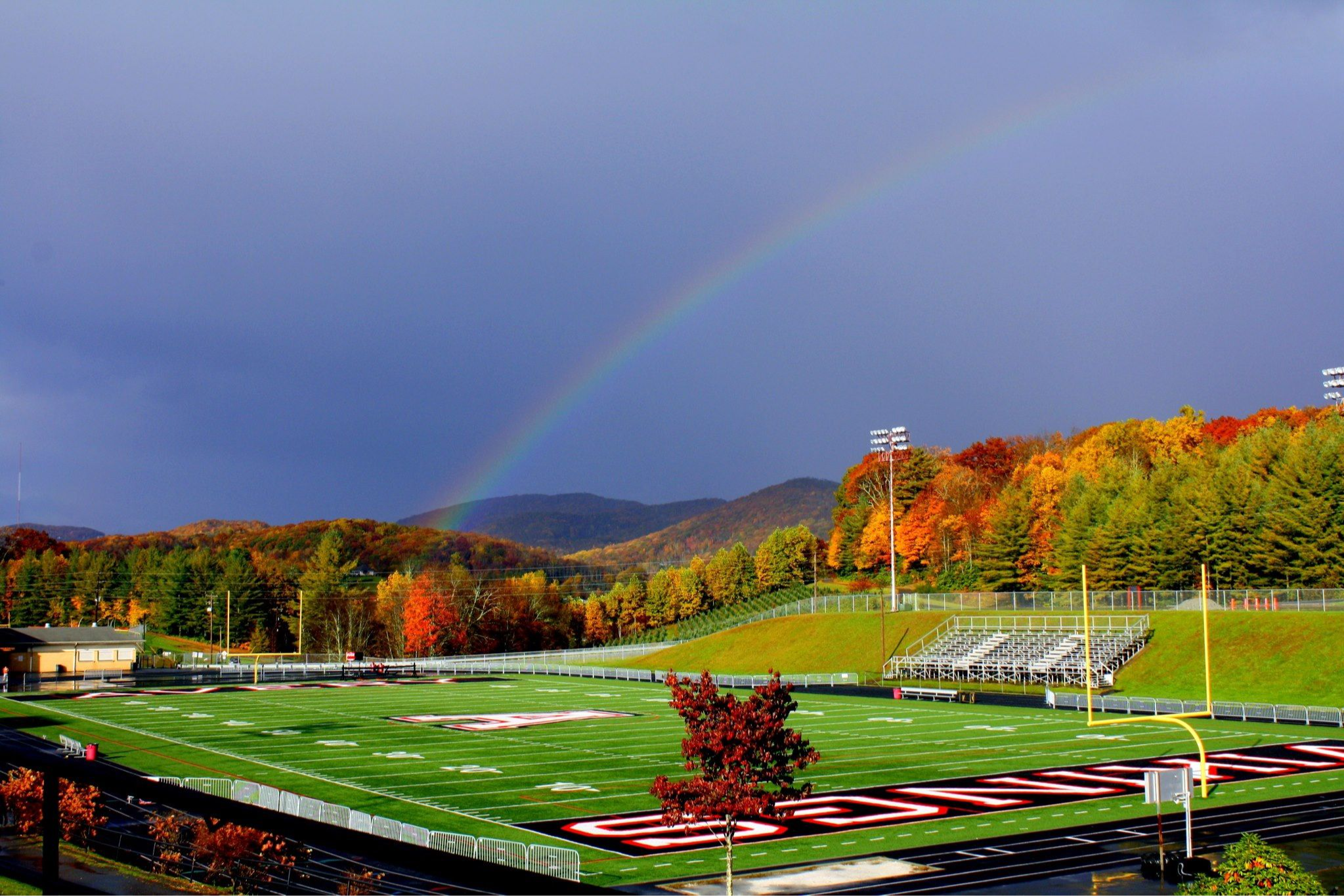 A distant view of the football field with colorful leaves on the trees, dark skies and a rainbow in the background.