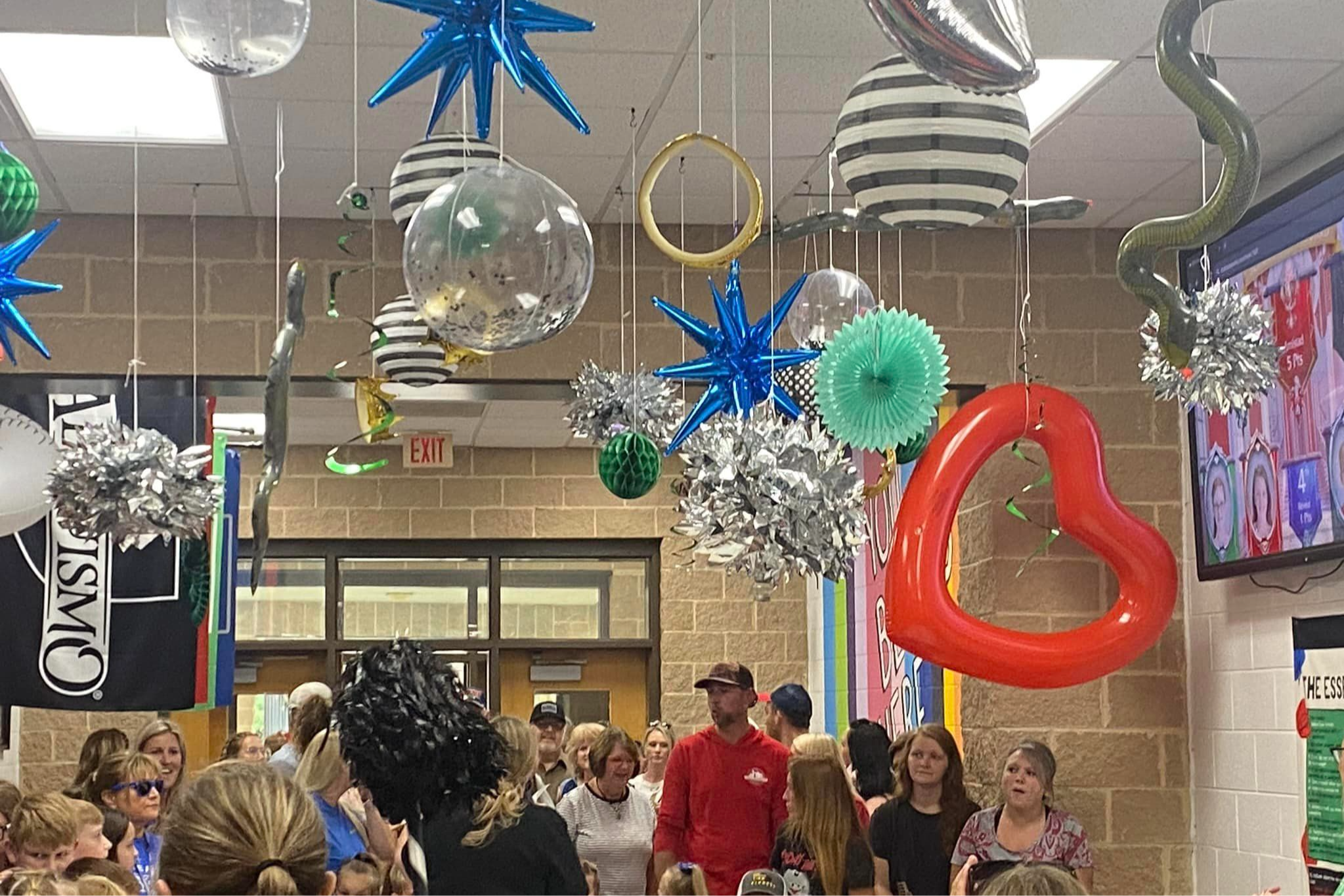 Main entrance hallway with a group of students, teachers and parents.  Silver pom poms, blue metallic stars, an inflated red hear balloon and other decorations are hanging from the ceiling.