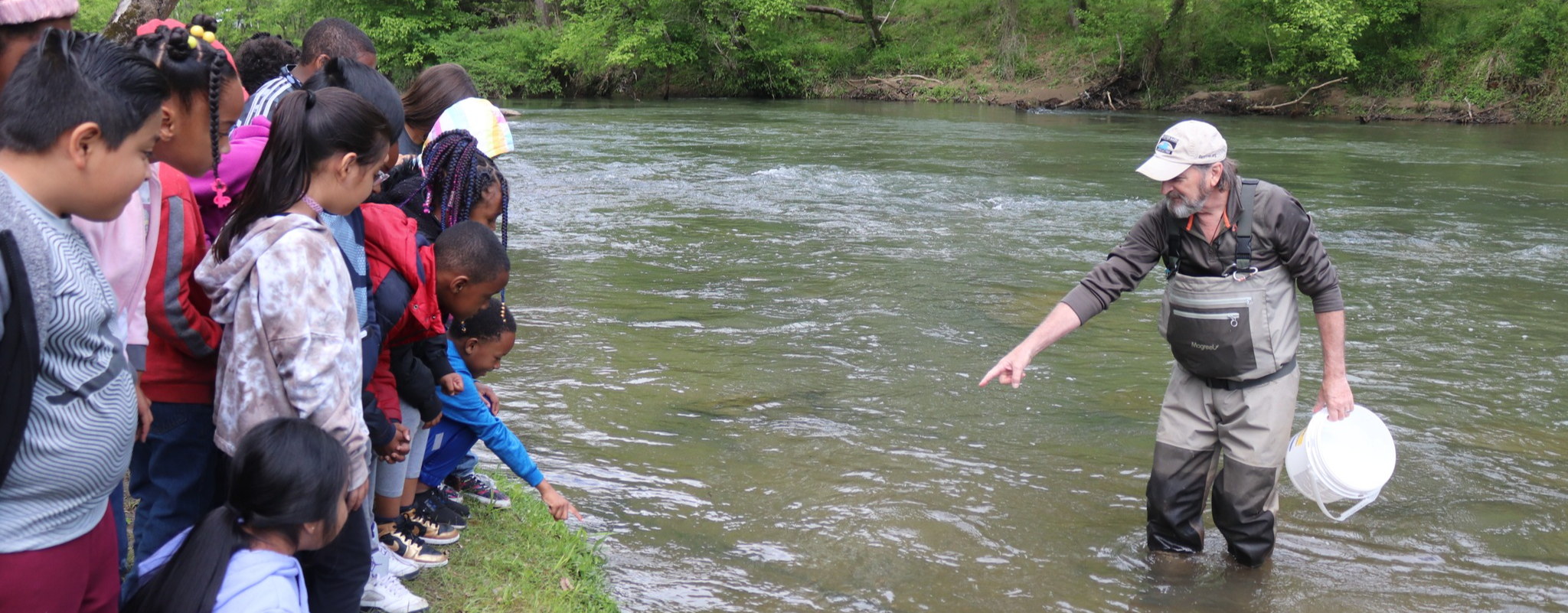 Students stand on a riverbank as a DRBA employee stands in the river and points to the water