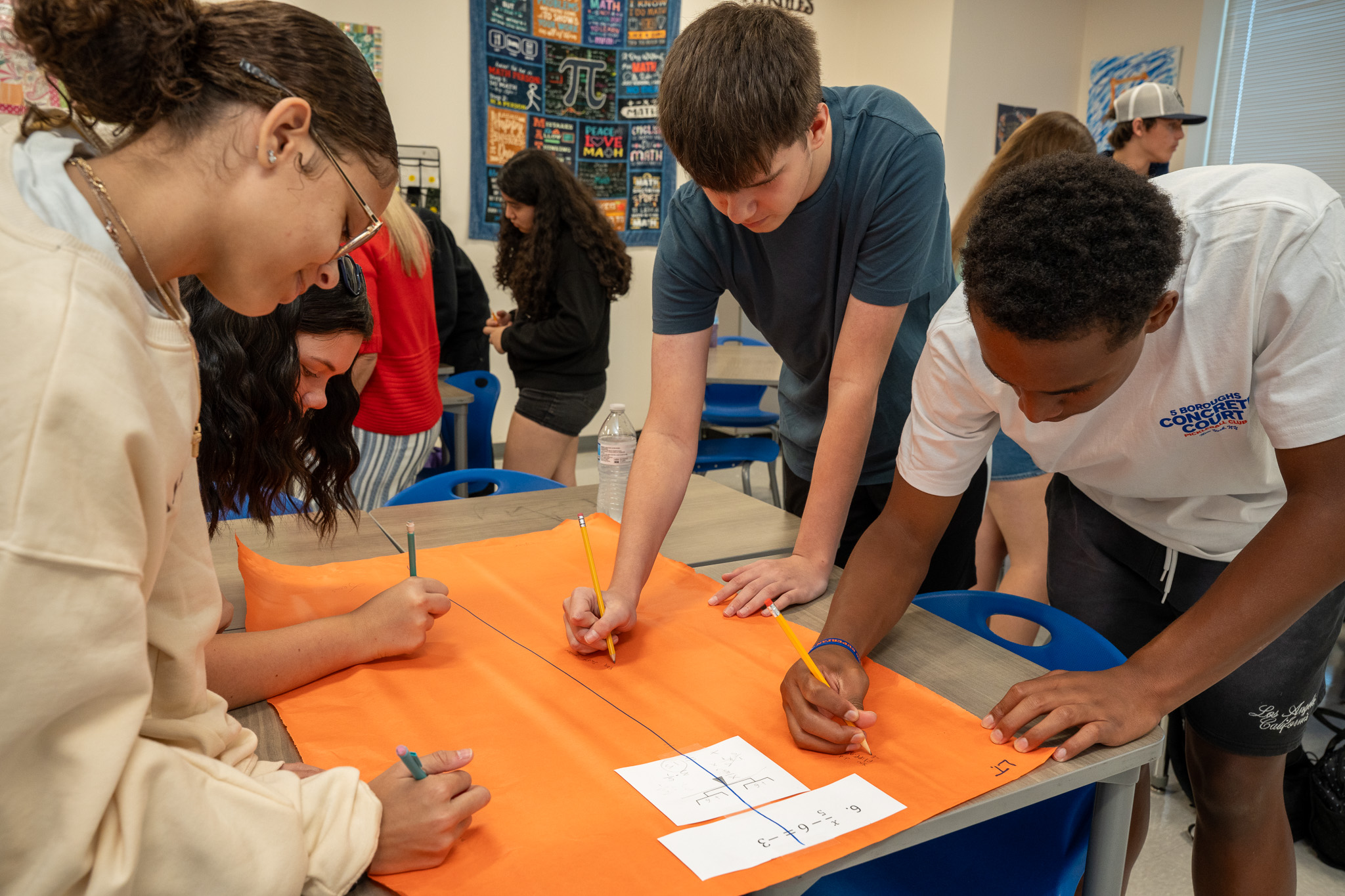 Four students collaborate around a desk in a math classroom, working together on an orange poster board. They are using pencils and rulers to draw geometric shapes, with a small reference sheet visible on the desk. Math posters and educational materials are displayed on the classroom walls in the background.