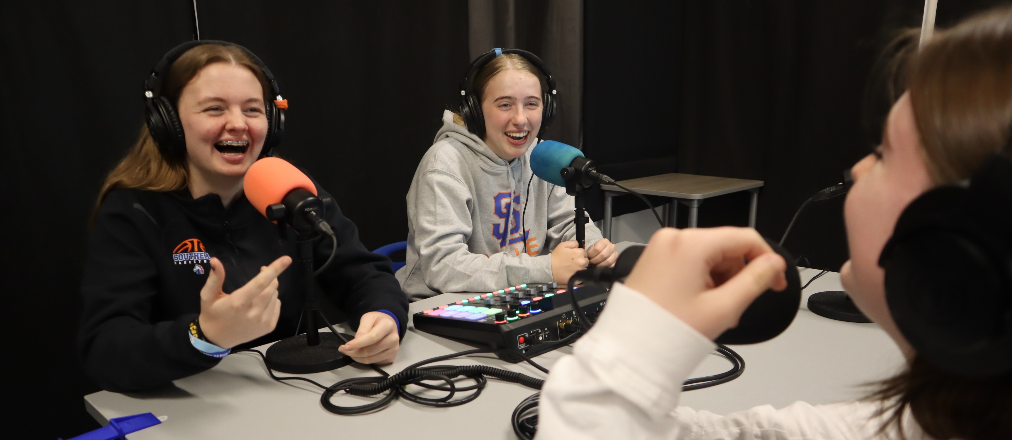 Three students sitting at a table in front of microphones, wearing headphones, laughing at a joke