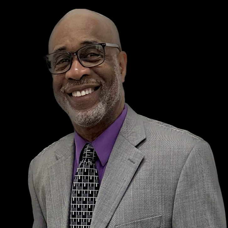 dark skinned man with glasses, bald head and a beard, wearing a gray suit jacket purple shirt and patterned tie. he is smiling