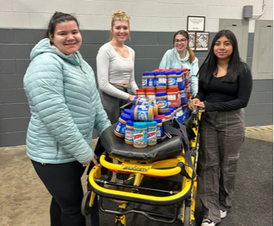 Four EMS students with a wheeled stretcher that is holding jars of peanut butter