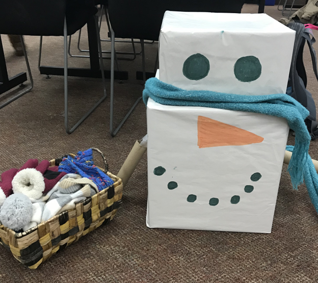 A snowman made out of two boxes with a basket of winter items to "dress" the snowman