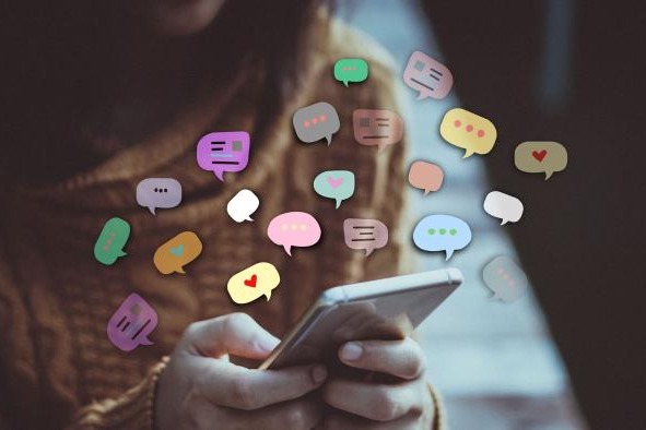 The image shows a person holding a smartphone, with various colorful speech bubbles floating around the device, symbolizing online conversations or social media interactions. The person's face is not visible, drawing attention to the smartphone and the digital messages.