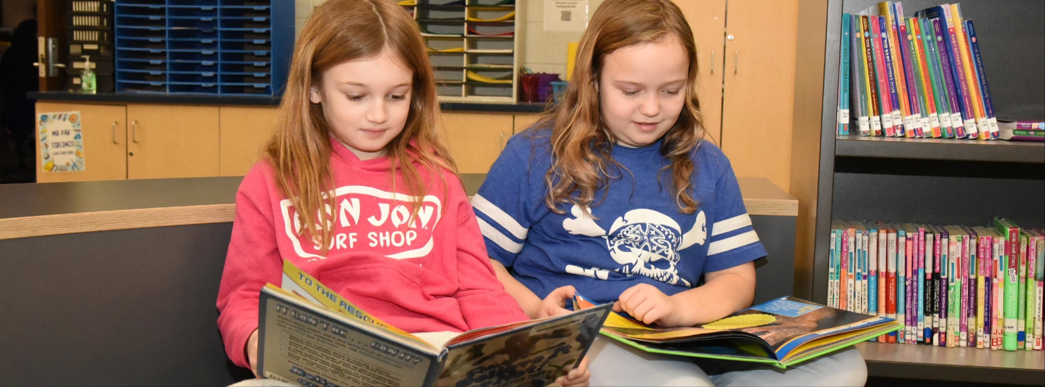 Two upper elementary students sit together in the school library, quietly reading their books.