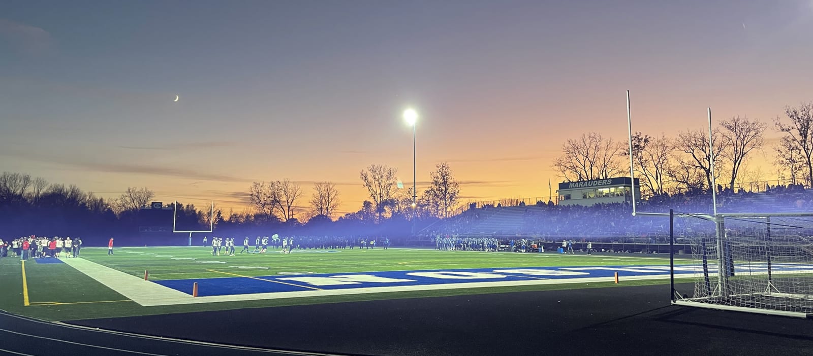 Football field photo taken from a distance during a game while the sun sets in the back and some fog/smoke rolls across the field