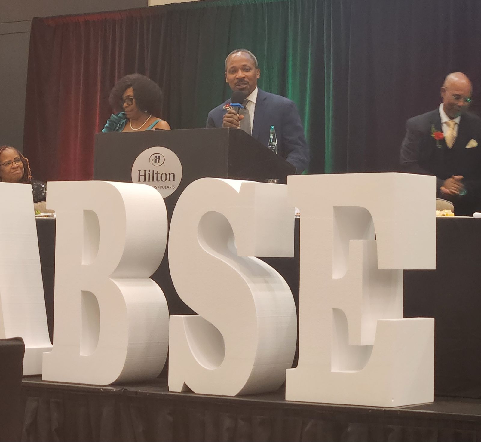 Dr. Jamison accepting his honor at the podium, behind large white letters that spelled out OABSE - not all the letters are visible in the photo.