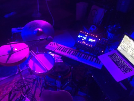A small drumkit, a piano, and a laptop with purple light shining on them