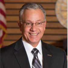 white male with grey hair & glasses wearing a dark suit with white shirt and grey striped tie