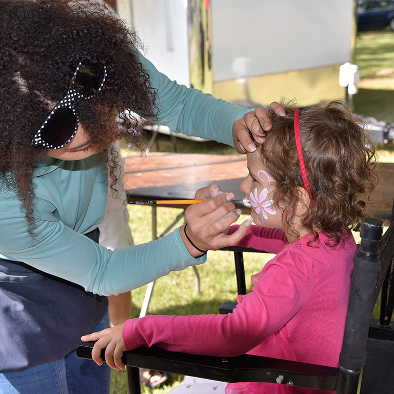 white female face painting a small female child in chair