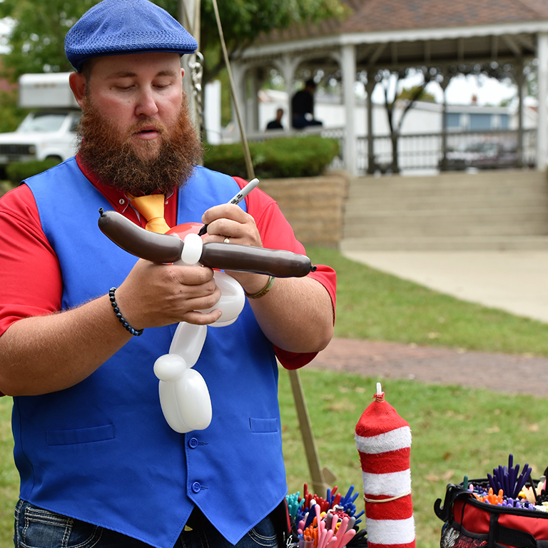 white male with beard wearing a blue hat and vest & red shirt making balloon animals