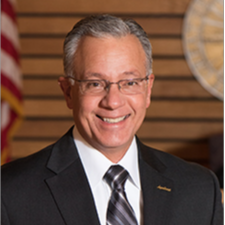 white male with grey hair & glasses wearing a dark suit with white shirt and grey striped tie